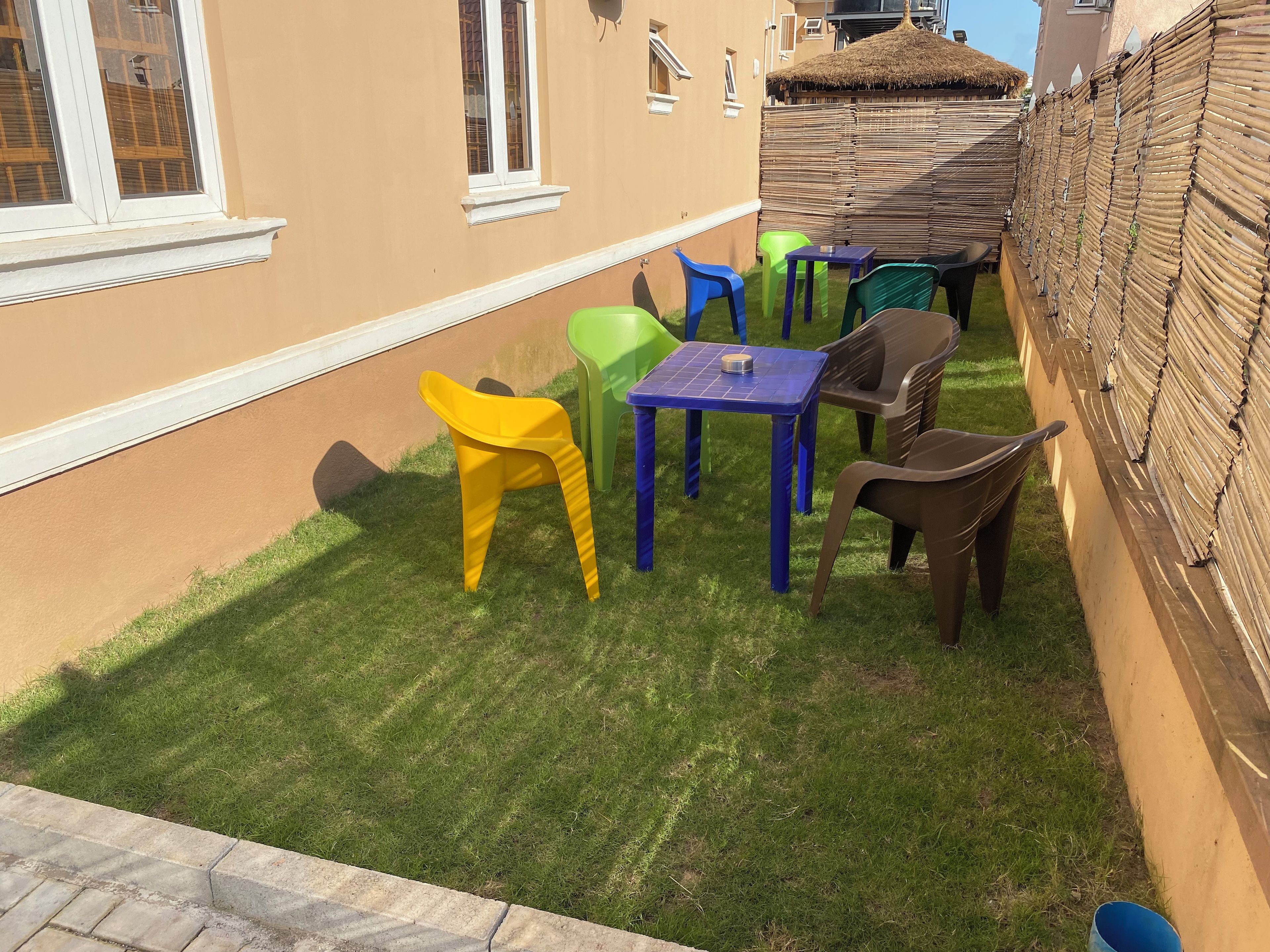 A small outdoor seating area with plastic chairs and tables, a thatched roof structure, and a bamboo fence.