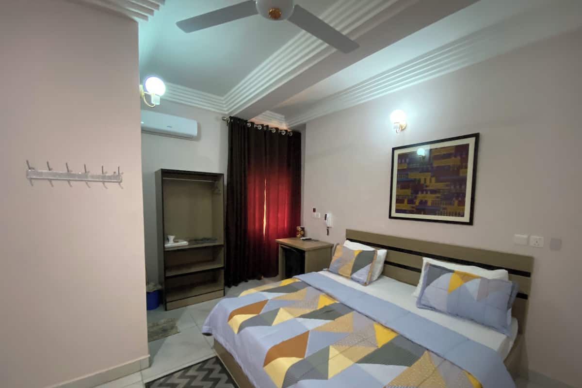 A bedroom with a bed, a ceiling fan, an air conditioning unit, a nightstand, a lamp, a framed picture, and a wardrobe.