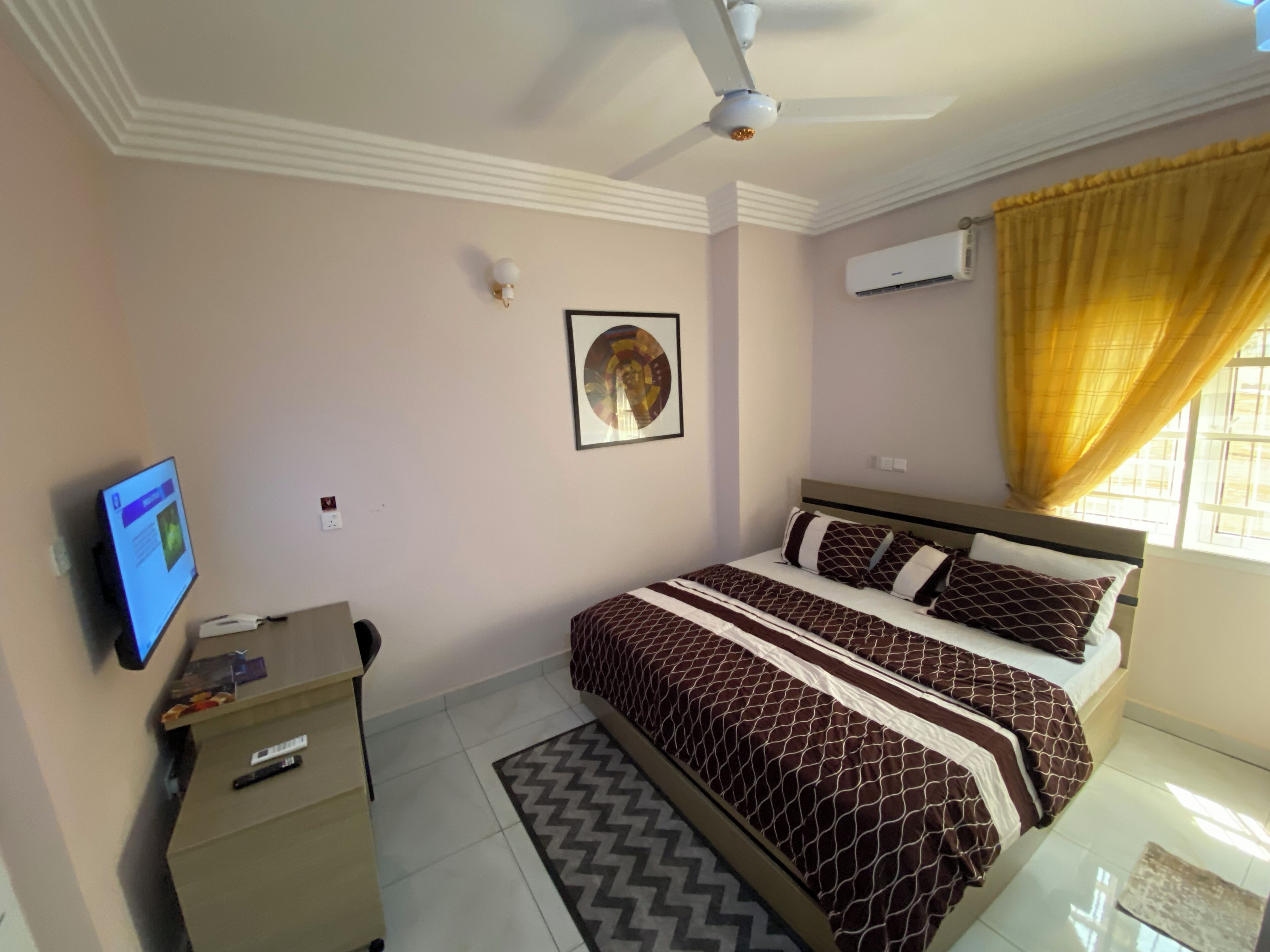A bedroom with a bed, a desk, a ceiling fan, an air conditioner, and a window with curtains.