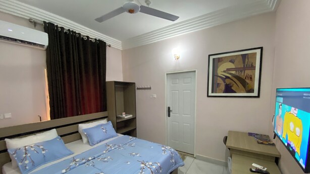 A bedroom with a bed, a desk, a television, a door, and an air conditioning unit. - C7 Hazelwood Residence & Suites (Abuja)