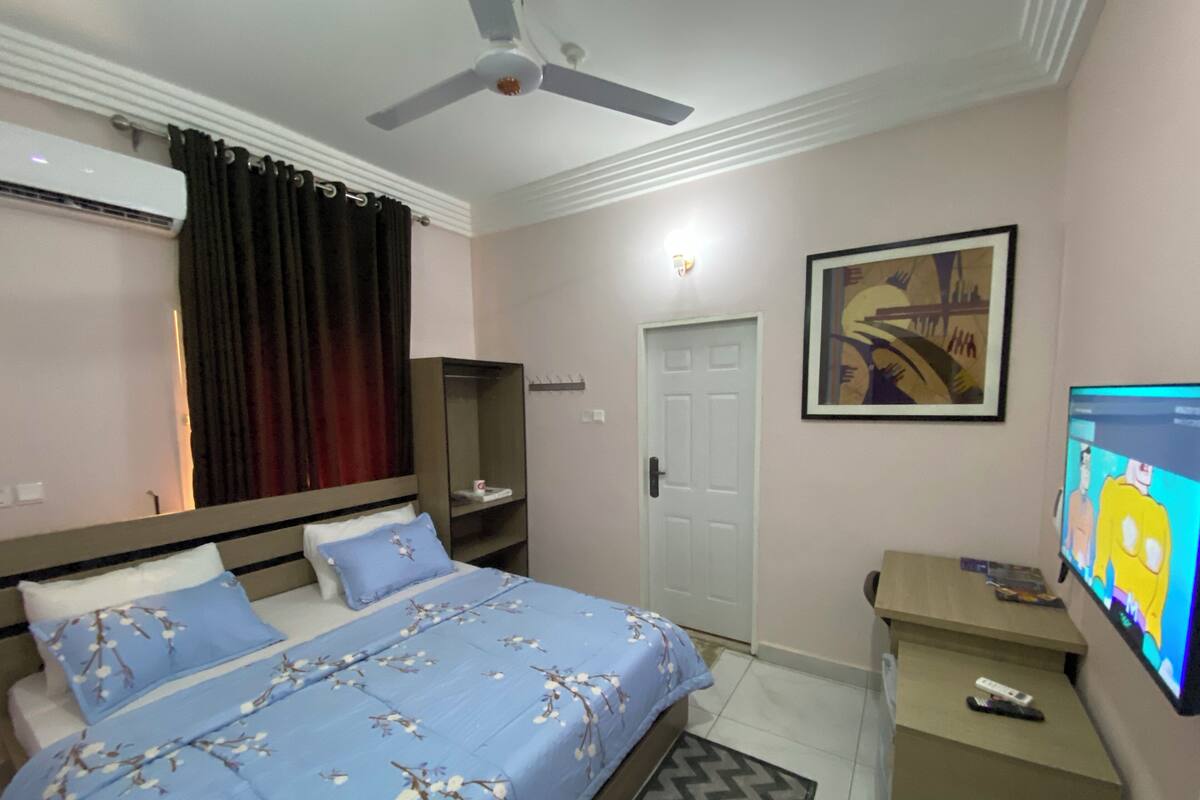 A bedroom with a bed, a desk, a television, a door, and an air conditioning unit.