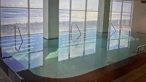 An indoor swimming pool with clear glass walls, metal handrails, and a tiled floor.
