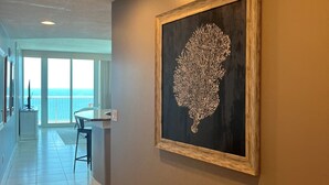 A hallway with a framed artwork on the wall, a dining area with a view to the outside, and a tiled floor.