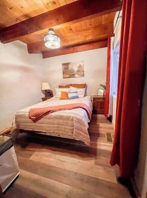 Quarto com teto de madeira, cama com roupa de cama listrada, mesa de cabeceira, luminária e pintura na parede.