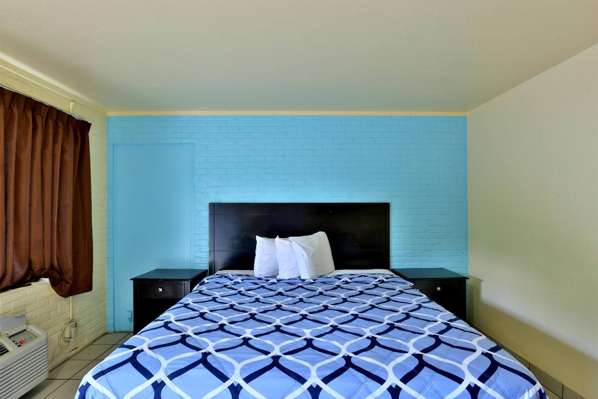 Ein Hotelzimmer mit blauer Ziegelwand, einem Bett mit geometrischem Muster auf der Bettdecke, zwei weißen Kissen, einem schwarzen Kopfteil und einem kleinen Nachttisch.