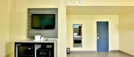 Habitación de hotel con televisión, kitchenette, dos camas, un espejo y una puerta que da al pasillo.