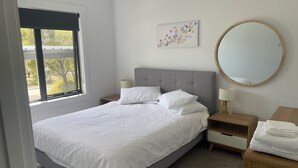 A neatly made bed with white linens, a grey upholstered headboard, and a bedside table with a lamp. A round mirror hangs on the wall above the bed. - Little White House on Ecclestone close to city centre and Tamar Valley Wineries! (Riverside)