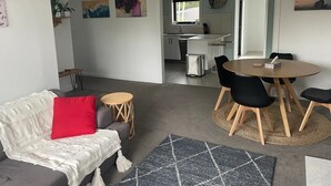 A modern living room with a sofa, a dining table with chairs, a kitchen in the background, and a view of the outdoors. - Little White House on Ecclestone close to city centre and Tamar Valley Wineries! (Riverside)