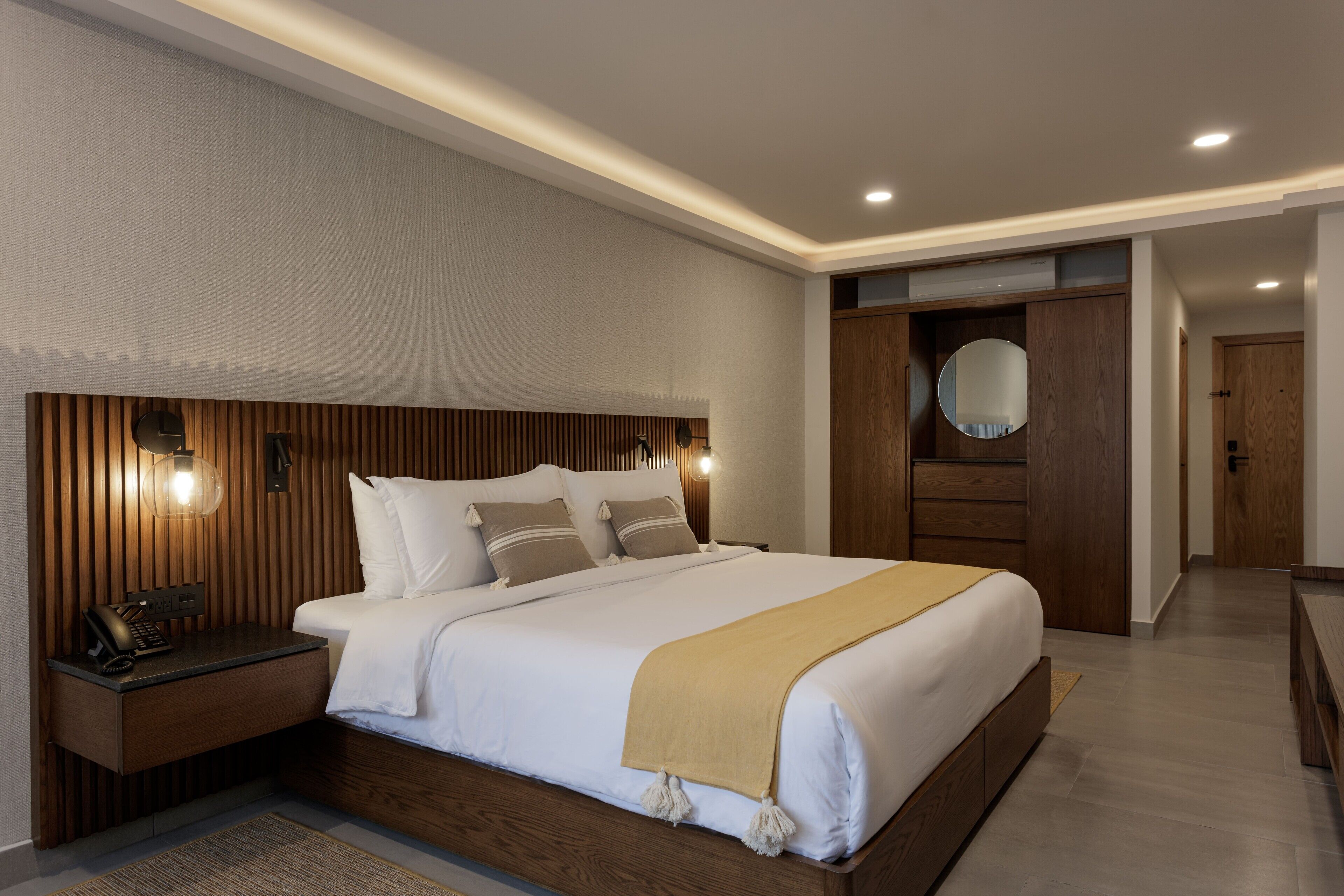 A modern bedroom with a wooden headboard, a bedside table with a lamp, and a large mirror above the bed.