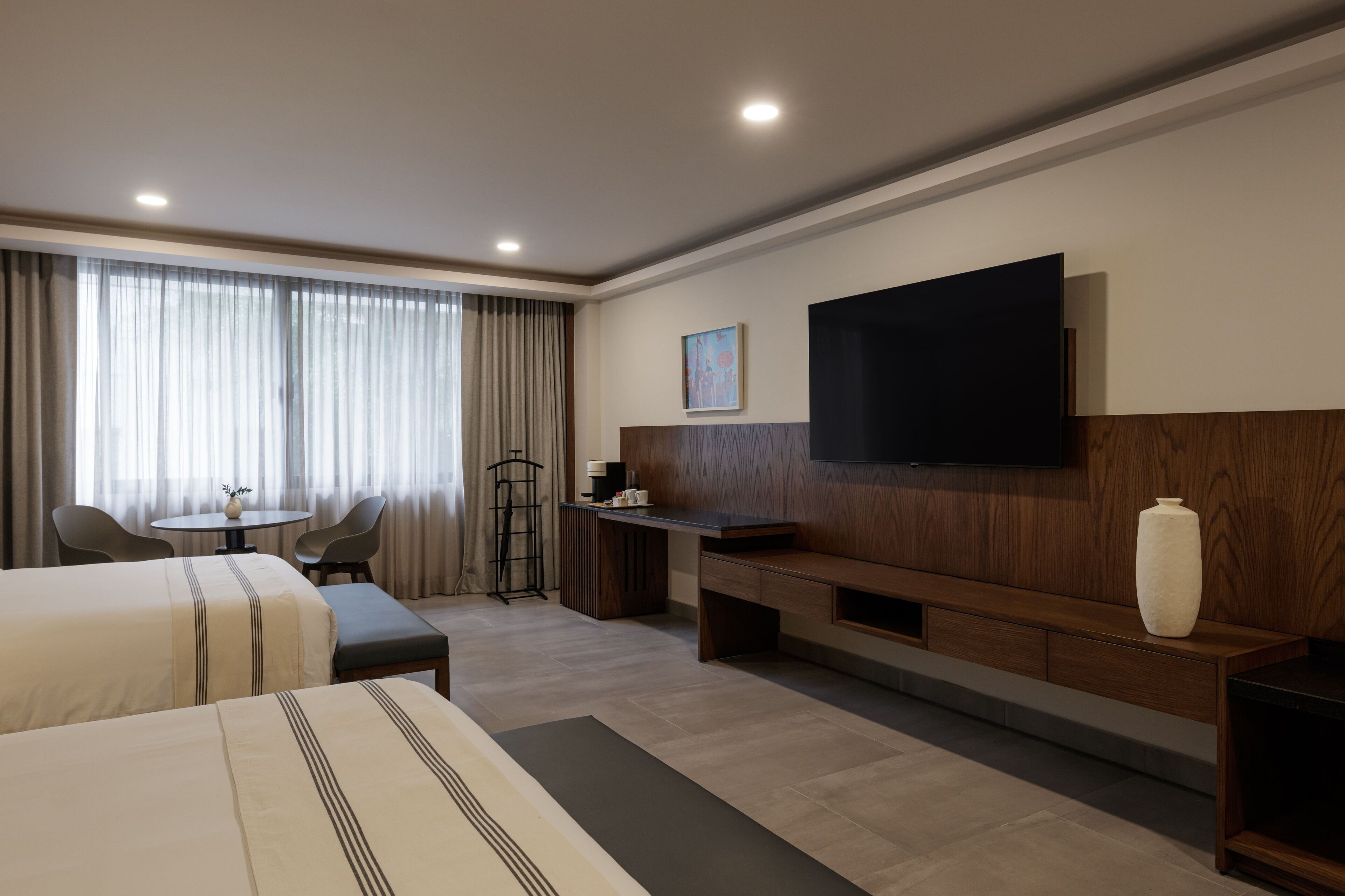 Habitación de hotel con dos camas, un escritorio, una silla, un televisor montado en la pared y un ventanal con cortinas.