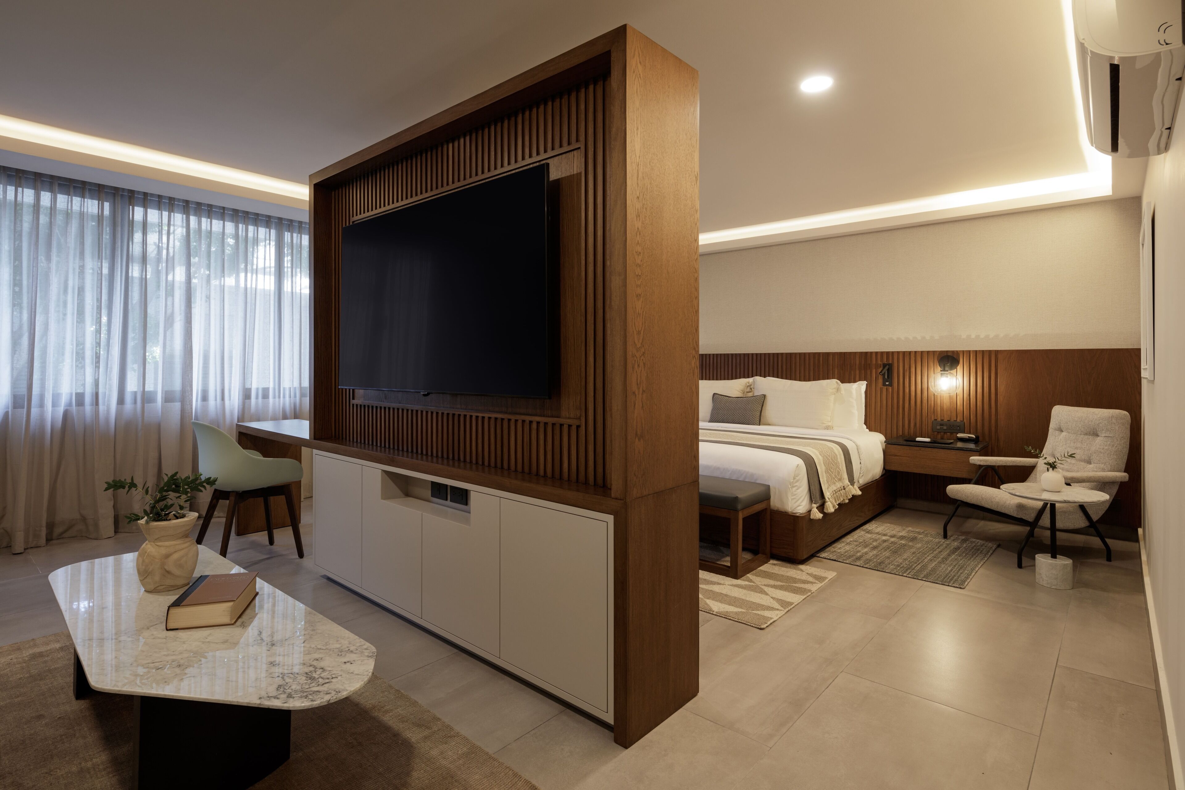 Habitación de hotel moderna con un televisor de pantalla plana grande, una cama con cabecera de madera, una zona de estar con una silla y una mesita, y una planta en maceta.