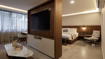 Habitación de hotel moderna con un televisor de pantalla plana grande, una cama con cabecera de madera, una zona de estar con una silla y una mesita, y una planta en maceta.
