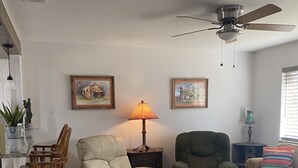 A living room with a beige sofa, a dark green armchair, a wooden chair, a floor lamp, and two framed paintings on the wall.