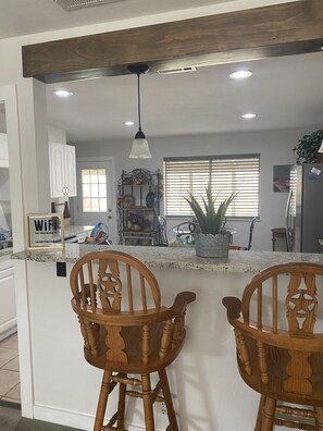 Uma cozinha com ilha central, cadeiras de madeira, luminária pendente e janela com persianas.
