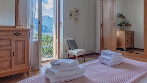 A room with a bed, a chair, a wooden dresser, and a large mirror. There is a balcony with a view of mountains. - Villa Gard by Wonderful Italy (Bellagio)