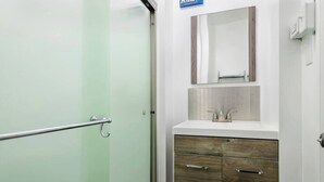 A bathroom with a glass door, a white sink with a mirror above it, and wooden cabinets below. - Modern Beachfront Suite 301 (Ocean City)