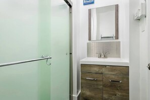 Un baño con puerta de vidrio, un lavabo blanco con espejo y armarios de madera debajo.