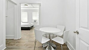 Ein heller Raum mit einem runden Tisch, vier Stühlen, einem Bett im Hintergrund und einer Tür rechts.