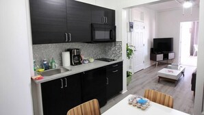 Une cuisine moderne avec des meubles de couleur sombre, un four à micro-ondes et une machine à café. On y trouve un coin repas avec une table et des chaises blanches. On aperçoit un téléviseur à l’arrière-plan.