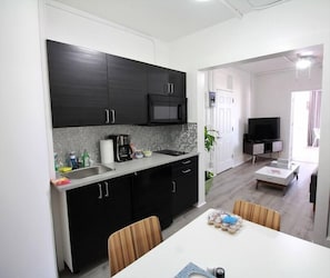 A modern kitchen with dark cabinets, a microwave, and a coffee maker. There is a dining area with a white table and chairs. A TV is visible in the background. - Modern Beachfront Suite 201 (Ocean City)