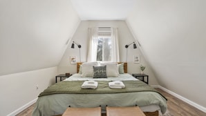 Ein Schlafzimmer mit schräg versiegelter Decke, einem Bett mit grüner Bettdecke, zwei Nachttischen mit Lampen, einem Fenster mit Vorhängen und einer Bank am Fußende des Bettes.