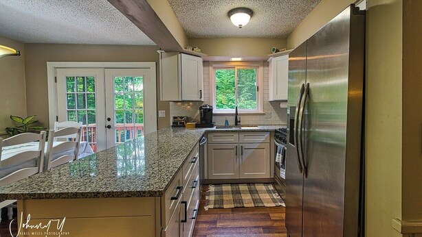 Una cocina con encimeras de granito, armarios blancos, un refrigerador de acero inoxidable y una ventana con vistas a la vegetación.