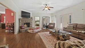 A spacious living room with hardwood flooring, a stone fireplace, a ceiling fan, and a large flat-screen TV mounted on the wall. - Summer Snow: Large Pet-Friendly Family Home w Yard, Firepit | 6 Min to Downtown (Highlands)