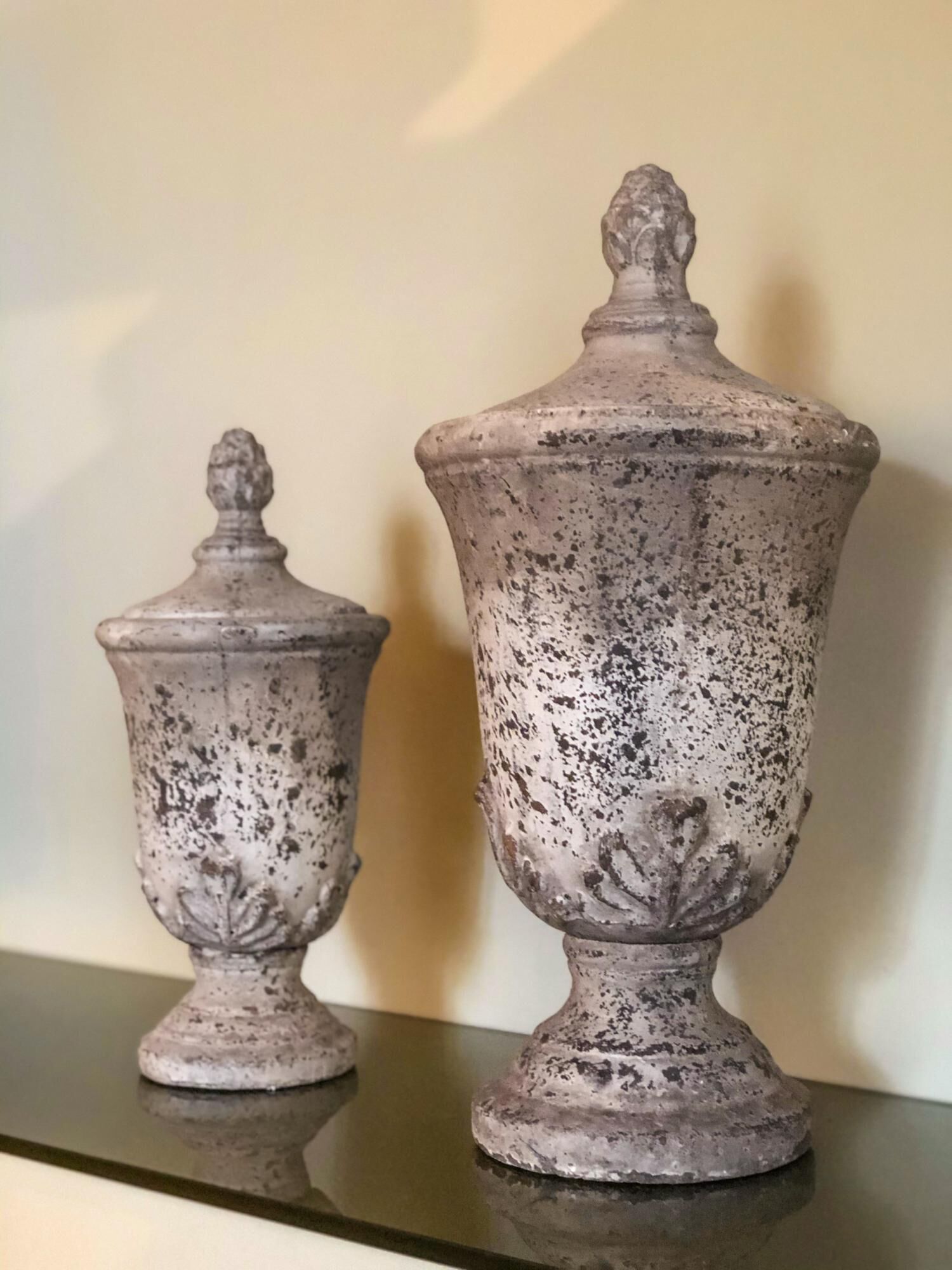 Two antique urns with a weathered finish, placed on a reflective surface against a plain wall.