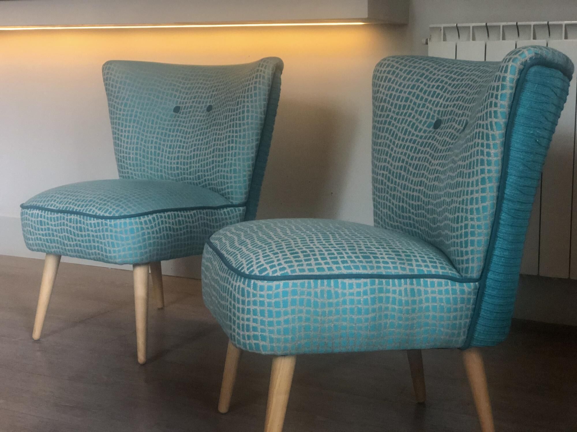 Twee teal gekleurde fauteuils met houten poten, geplaatst tegen een muur met een lichtbron erboven.