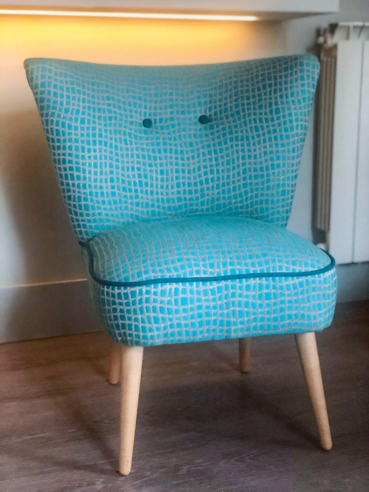 A teal patterned armchair with wooden legs, positioned in a room with a wooden floor and a white wall.