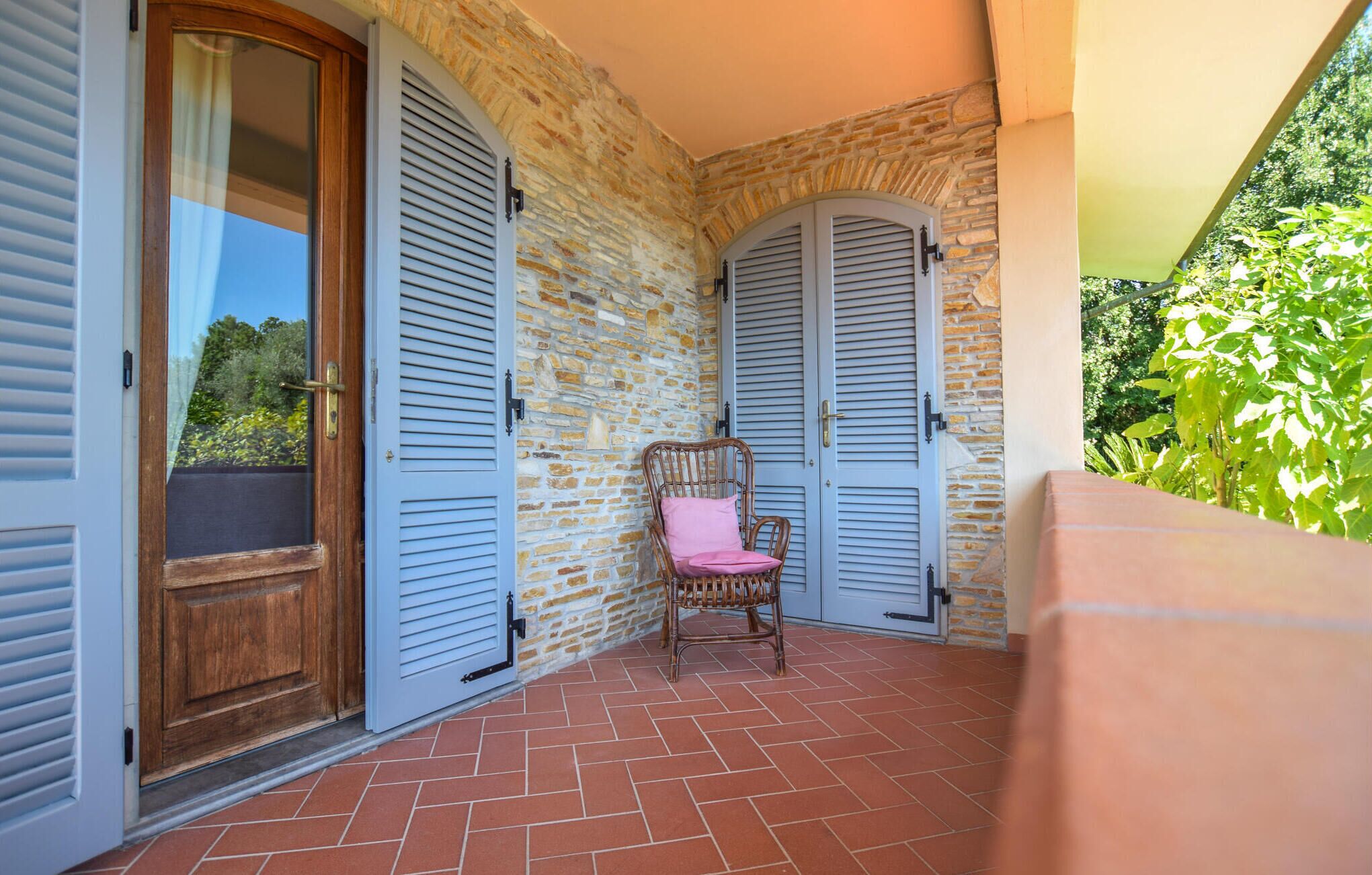 A balcony with stone walls, two blue shutters, a wicker chair with a pink cushion, and a wooden door.