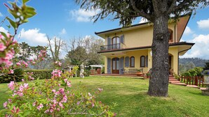 Une maison à deux étages dotée d’un balcon, entourée d’un jardin bien entretenu et d’un grand arbre.