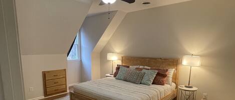 Un dormitorio con cama, ventilador de techo, mesita de noche, lámpara y ventana.