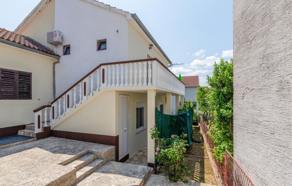 A two-story building with a white exterior, a staircase with white balusters, and a small garden area with a green fence and pink flowers. - 1 bedroom beautiful apartment in Karin Gornji (Karin Gornji)