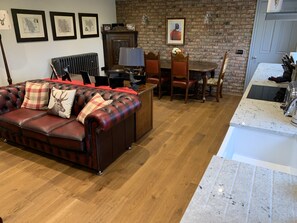 A cozy living room with a leather sofa, a dining area with a brick wall, and a kitchen with a white countertop.