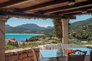 Uma mesa com tampo de vidro e cadeiras de vime, com vista para uma paisagem costeira com montanhas e uma praia.