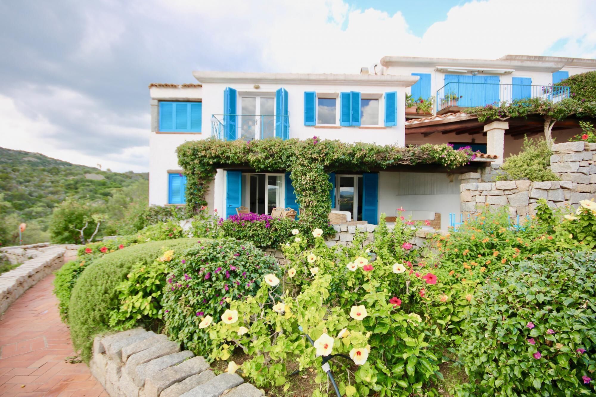 A two-story building with blue shutters, a stone wall, and a garden with various flowers.