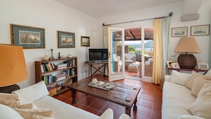 Un salon comprenant un canapé, une table basse, une bibliothèque, un téléviseur et une vue sur une terrasse extérieure, accessible par une porte vitrée.