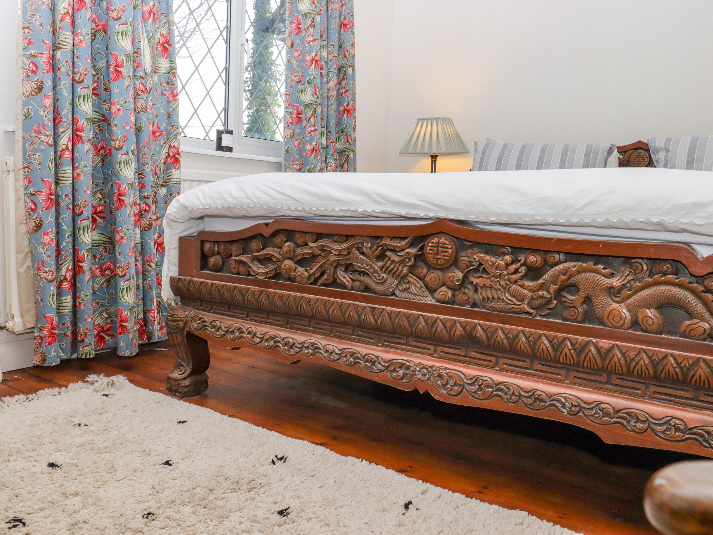 A bedroom with a wooden bed featuring intricate carvings, a fluffy white rug, and floral curtains.