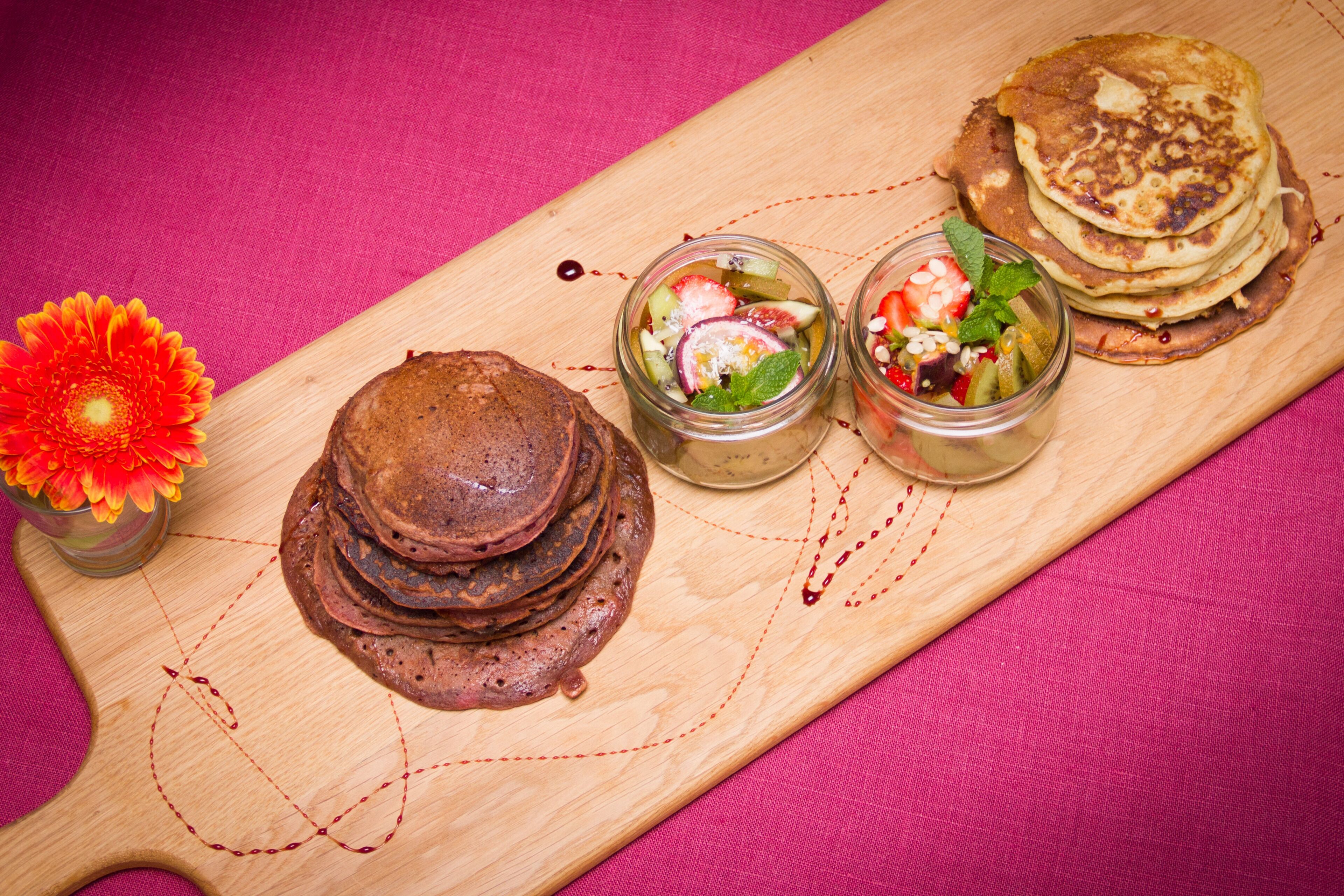 Een houten plank met pannenkoeken, een pot fruitsalade en een bloem.