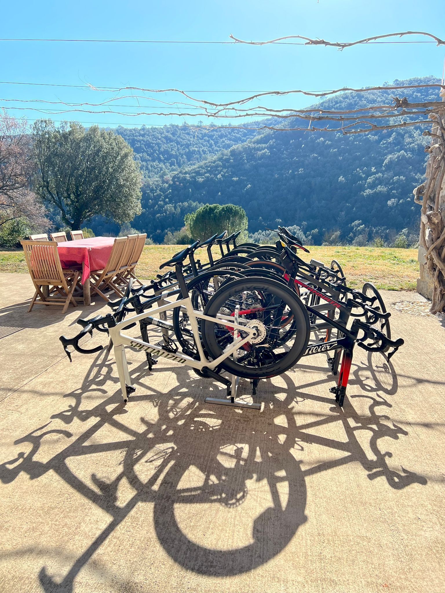 Een fietsenrek met meerdere fietsen geparkeerd op een betonvloer, een tafel met stoelen, en een bergachtig landschap op de achtergrond.