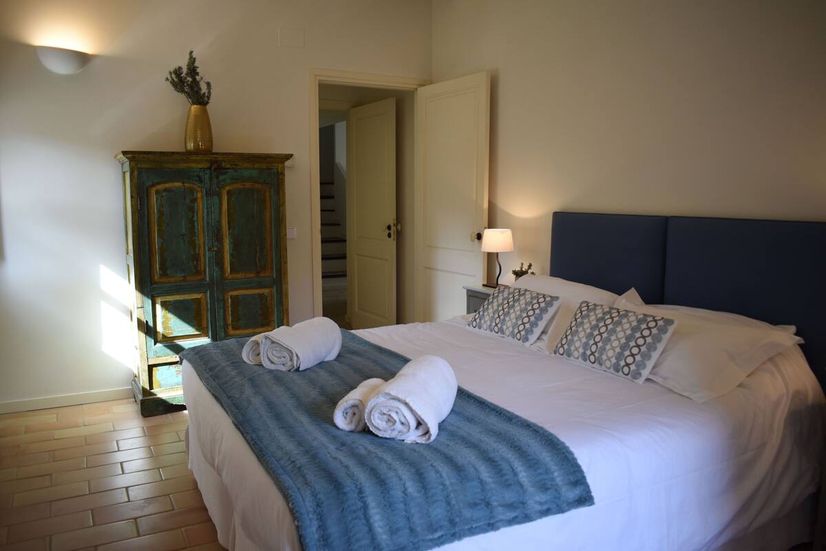 A neatly made bed with white linens and a blue blanket, a wooden cabinet with a vase, and a staircase in the background.
