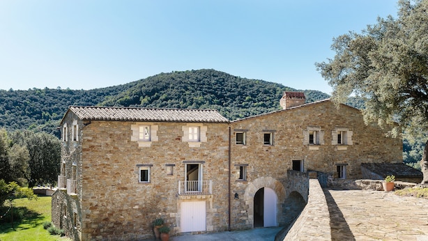 Un edificio de piedra con patio, entrada abovedada y vistas a una colina boscosa.