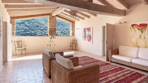 Amplia sala de estar con techo alto, vigas de madera, un ventanal con vista a un bosque, muebles de mimbre y un sofá blanco.