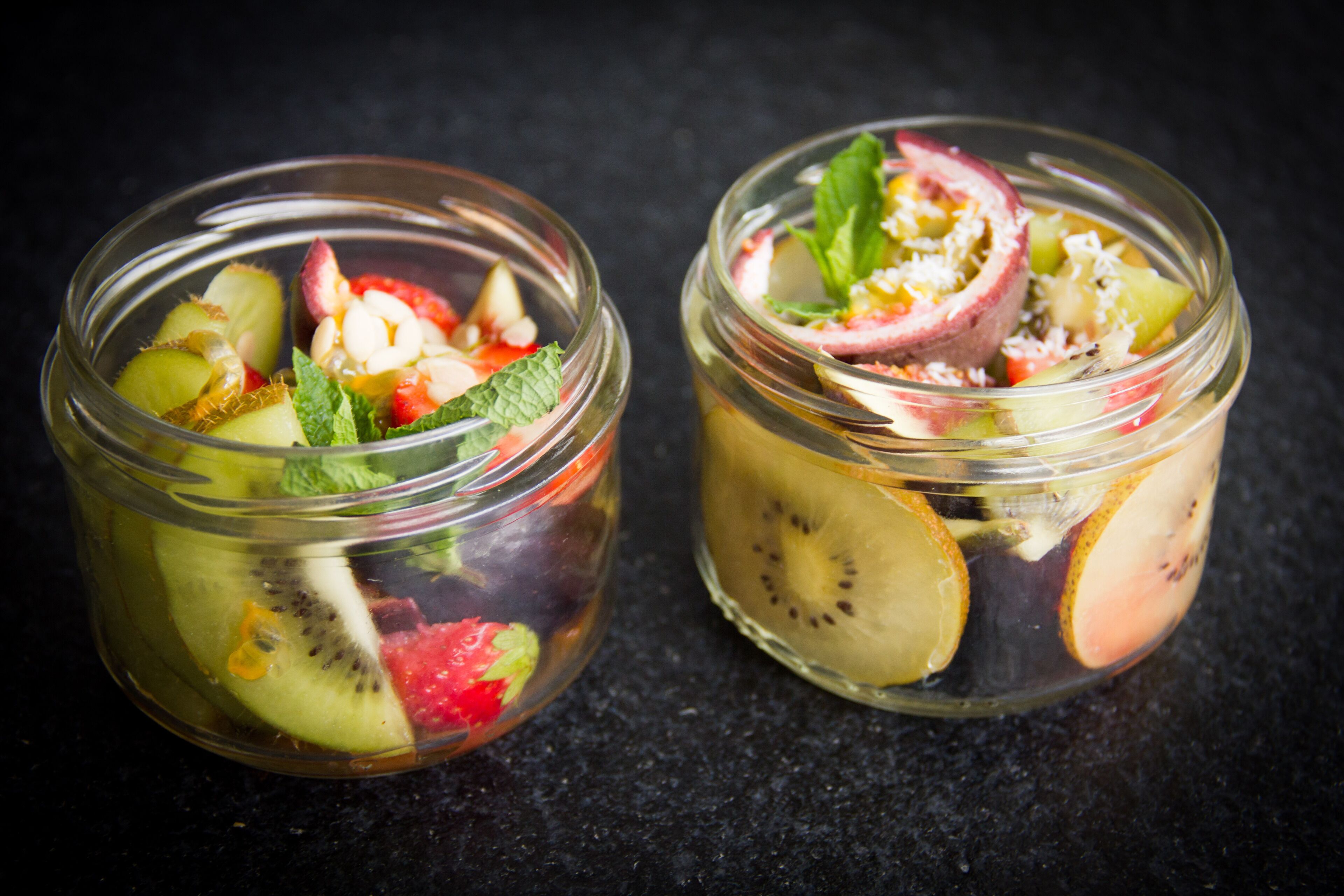 Deux bocaux en verre contenant une salade de fruits avec des tranches de kiwi, des fraises et des feuilles de menthe.