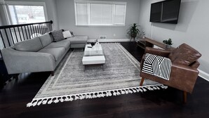 A modern living room with a grey sectional sofa, a coffee table, a patterned rug, a leather armchair, and a TV mounted on the wall. - Alexandria VA spacious, family-friendly 5BR Home near DC (Alexandria)