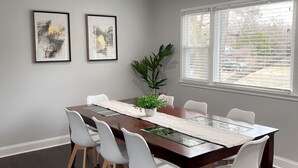 A dining room with a large table, six chairs, two framed artworks, a potted plant, and a window with blinds. - Alexandria VA spacious, family-friendly 5BR Home near DC (Alexandria)