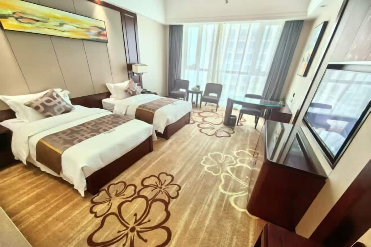 A hotel room with two beds, a TV, a desk, and a large window with curtains.