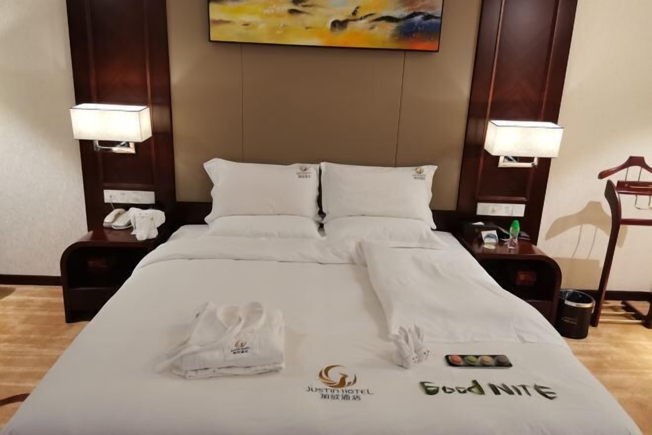 A hotel room with a neatly made bed, two bedside lamps, a painting on the wall, and a trash bin.
