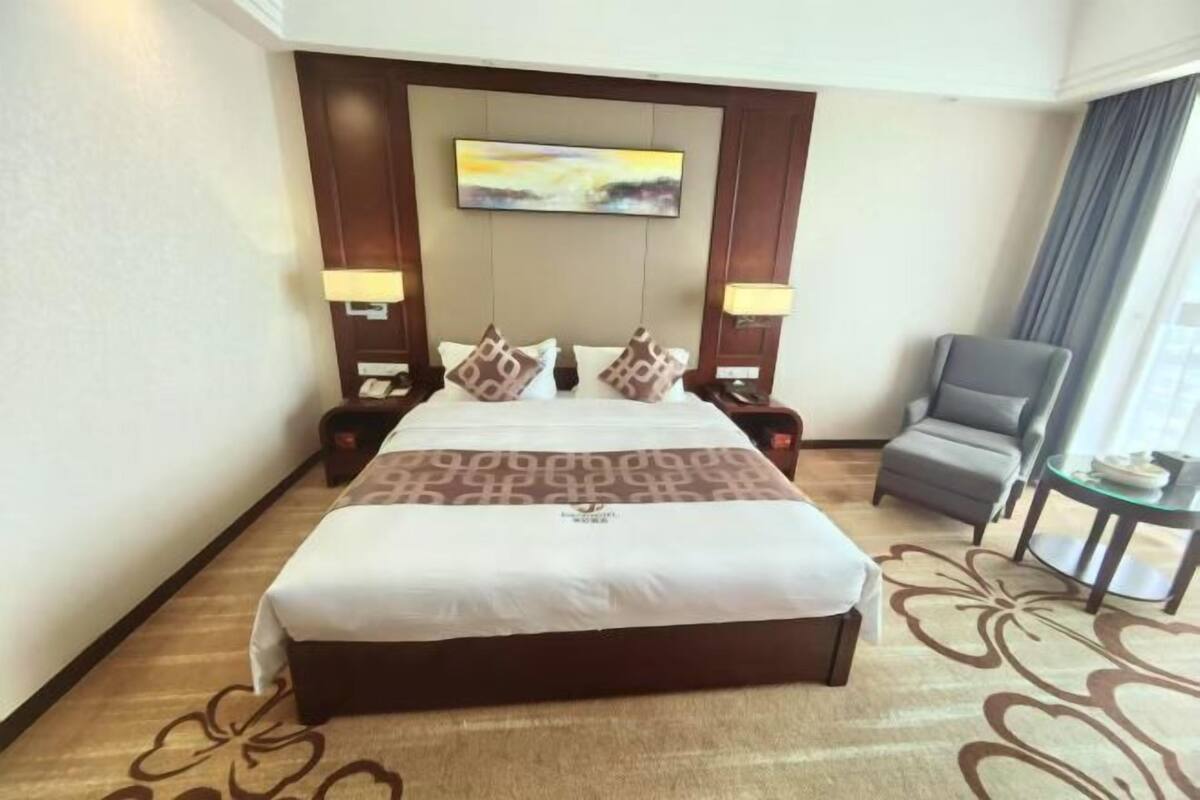 A hotel room with a large bed, two bedside tables with lamps, a gray armchair, and a small table with a plate.